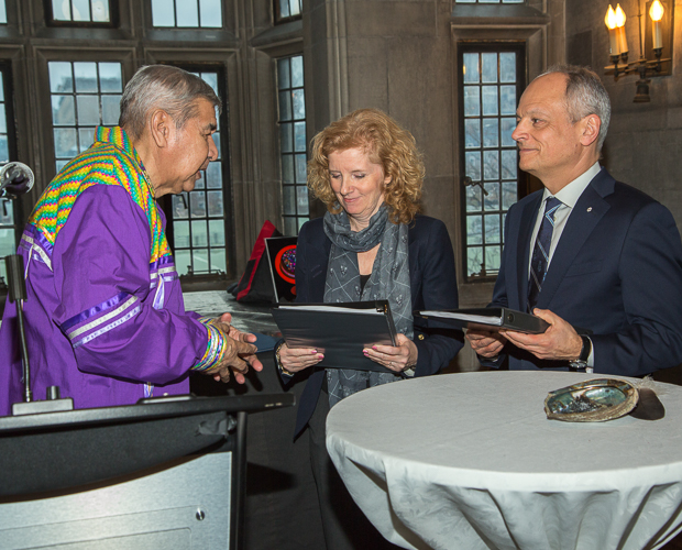 Meric Gertler holds a printed report and stands facing an Indigenous Elder, indoors at Hart House.