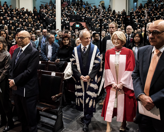 Meric Gertler and Rose Patten, both wearing academic robes, smile as they lead a procession into a crowded Convocation Hall.